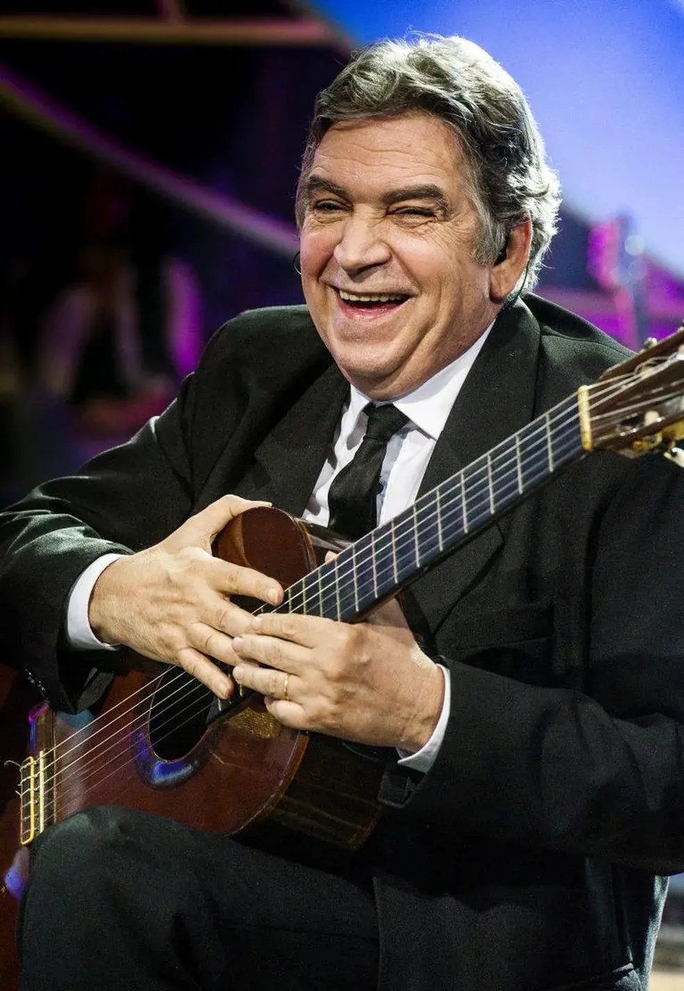 Produtor musical Guto Graça Mello sorri alegremente enquanto segura um violão clássico, vestindo um terno preto formal com gravata sobre uma camisa branca. A iluminação de palco destaca sua expressão vibrante em um cenário com tons de roxo e azul ao fundo.