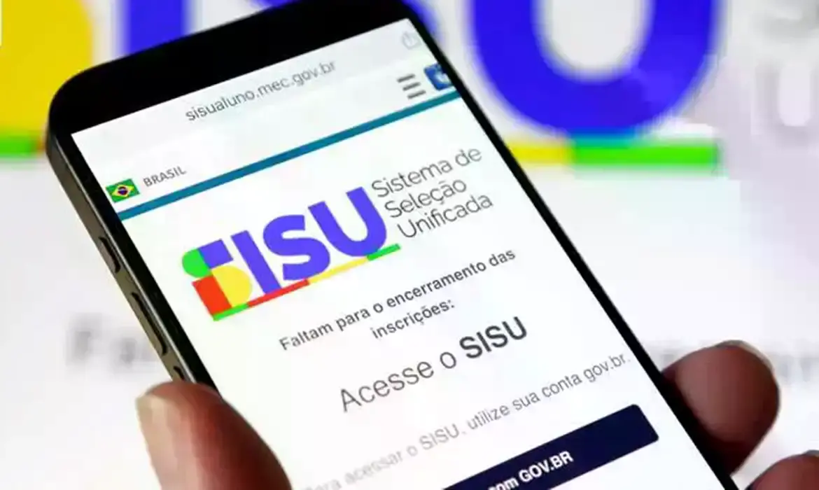 Um smartphone exibe a página oficial do Sistema de Seleção Unificada (SiSU), com o logotipo colorido e o texto 'Acesse o SISU' em destaque na tela. Ao fundo, o logotipo do programa aparece desfocado em um monitor de computador.