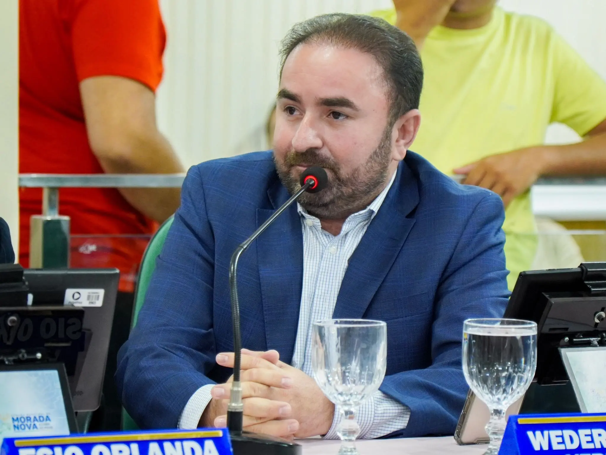 Homem de barba curta e cabelos escuros, usando paletó azul e camisa social clara, sentado em uma mesa de reunião com microfone à frente. Ele está com as mãos entrelaçadas, olhando para o lado, em ambiente que parece ser uma sessão legislativa ou audiência pública. Sobre a mesa há taças de água, monitores e placas de identificação. Ao fundo, aparecem pessoas desfocadas em pé.