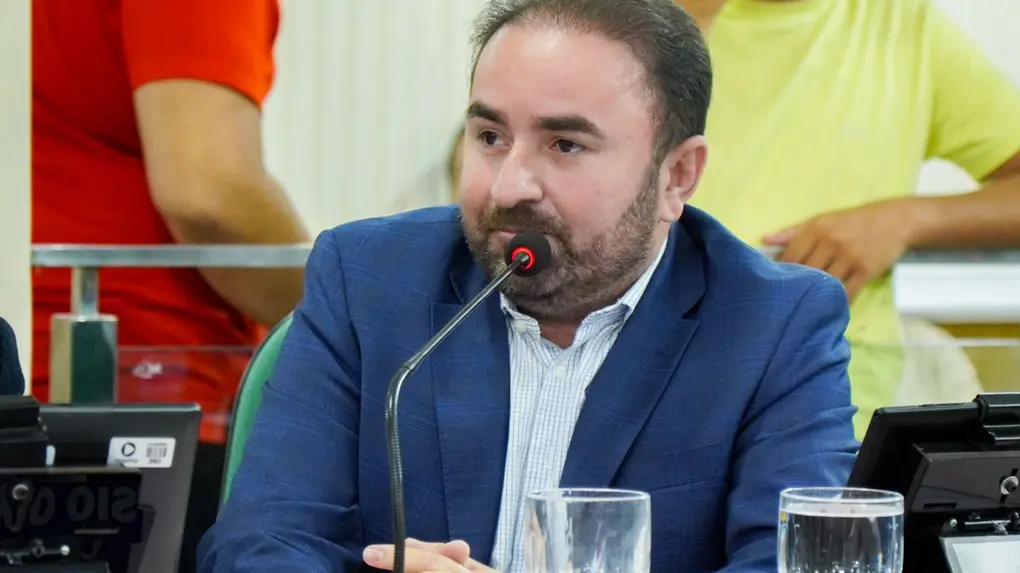 Homem de barba curta e cabelos escuros, usando paletó azul e camisa social clara, sentado em uma mesa de reunião com microfone à frente. Ele está com as mãos entrelaçadas, olhando para o lado, em ambiente que parece ser uma sessão legislativa ou audiência pública. Sobre a mesa há taças de água, monitores e placas de identificação. Ao fundo, aparecem pessoas desfocadas em pé.