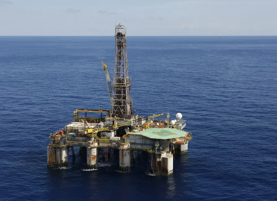 Imagem aérea de uma plataforma de petróleo em alto-mar, cercada por águas azuis calmas. A estrutura metálica semissubmersível ocupa o centro da imagem, com torre de perfuração elevada, equipamentos industriais e heliponto visível na parte superior. O horizonte do oceano aparece ao fundo sob céu claro com poucas nuvens.