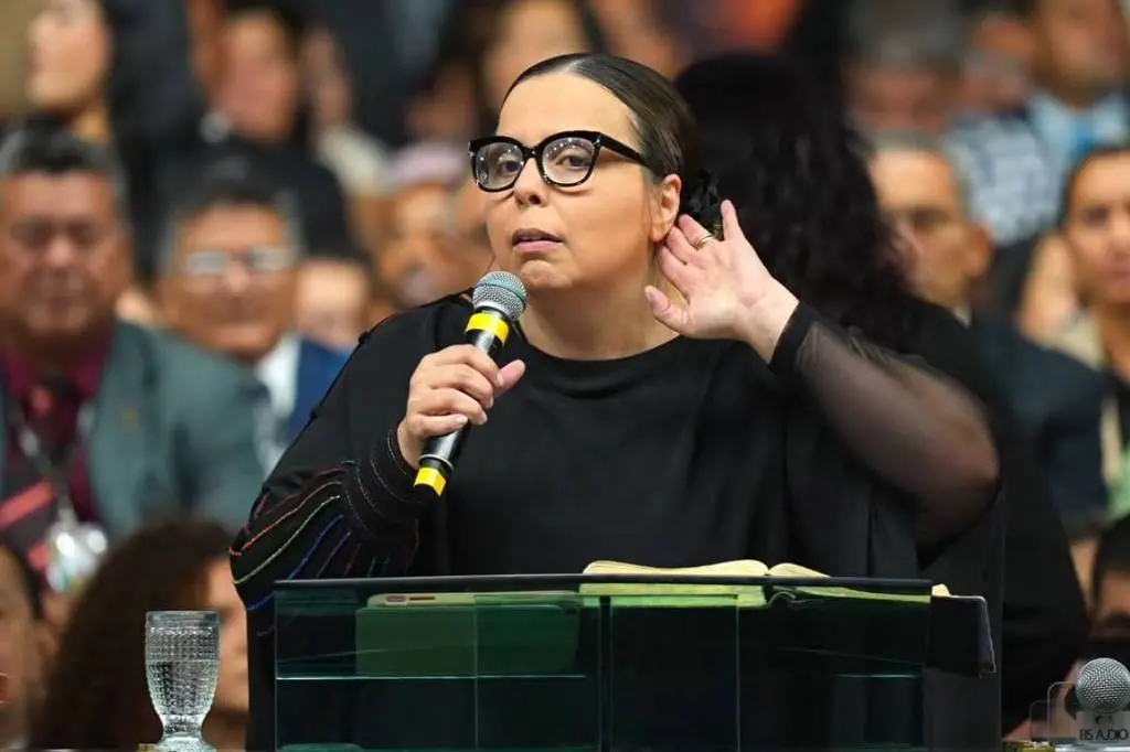 Mulher em um evento falando ao microfone, com uma pessoa ao fundo e uma plateia de espectadores atentos.