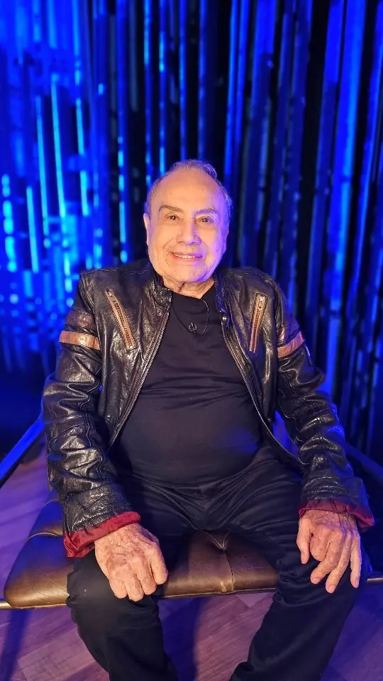 Homem idoso sorrindo, sentado em um palco com fundo de luzes azuis, usando jaqueta de couro e camiseta preta, em ambiente de evento musical ou teatral