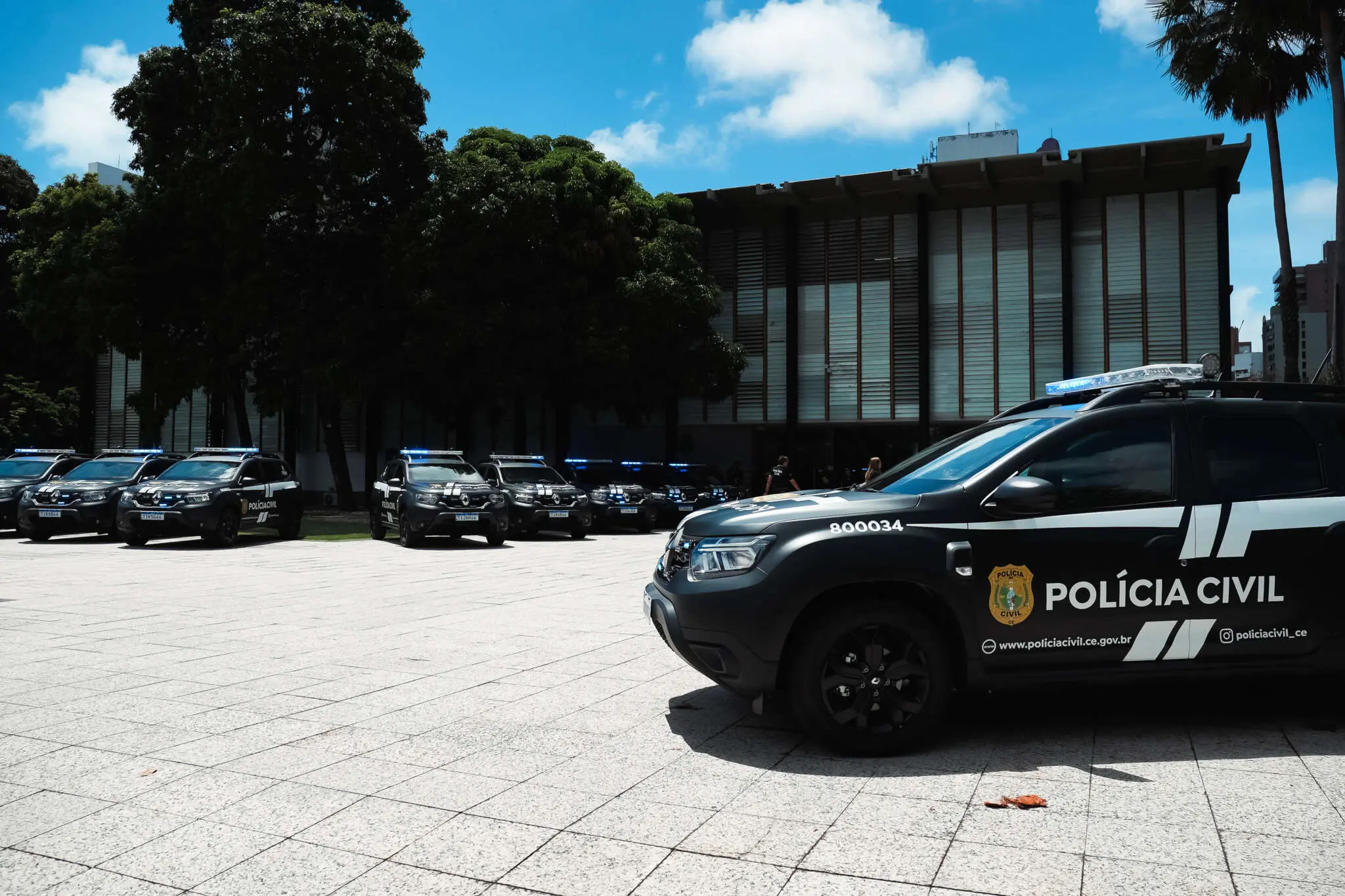Várias viaturas pretas da Polícia Civil do Ceará estão perfiladas em um pátio aberto, com destaque para um veículo em primeiro plano que exibe o brasão e a identificação oficial. Ao fundo, árvores densas e a fachada de um edifício governamental completam o cenário sob um céu azul ensolarado.