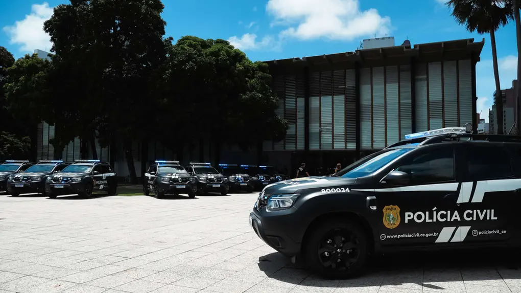 Várias viaturas pretas da Polícia Civil do Ceará estão perfiladas em um pátio aberto, com destaque para um veículo em primeiro plano que exibe o brasão e a identificação oficial. Ao fundo, árvores densas e a fachada de um edifício governamental completam o cenário sob um céu azul ensolarado.
