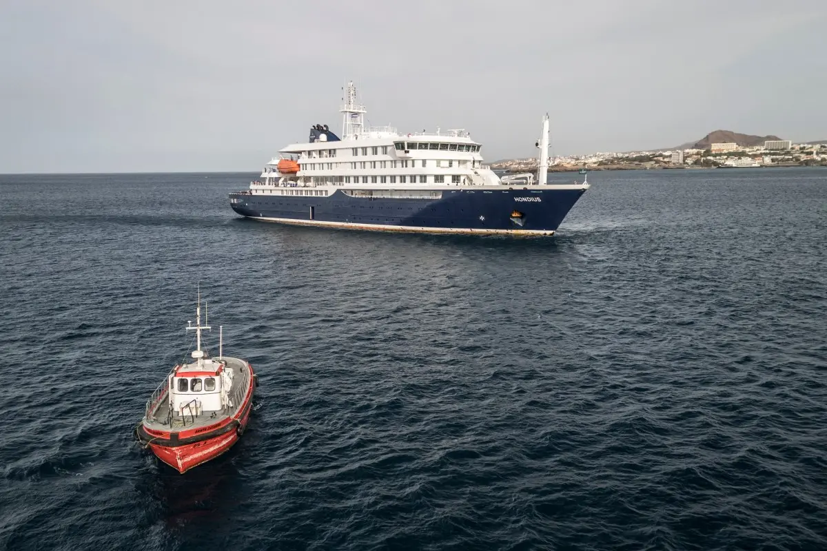 Navio de cruzeiro azul e branco navega em mar aberto, com pequena embarcação vermelha em primeiro plano e cidade costeira ao fundo.