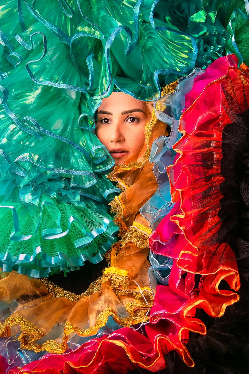 Rosto da rainha junina Mara Alexandre aparece centralizado e emoldurado por camadas de tecidos volumosos e coloridos em tons de verde, rosa, laranja e preto. A iluminação destaca a textura das fitas e babados que compõem o adereço ao redor da artista.