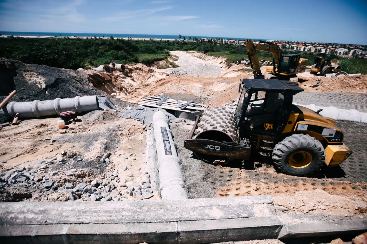 Canteiro de obras na rodovia com um rolo compressor amarelo em destaque sobre solo de terra e brita. Ao fundo, veem-se manilhas de concreto instaladas em uma vala, prédios residenciais e um outdoor sob céu aberto.