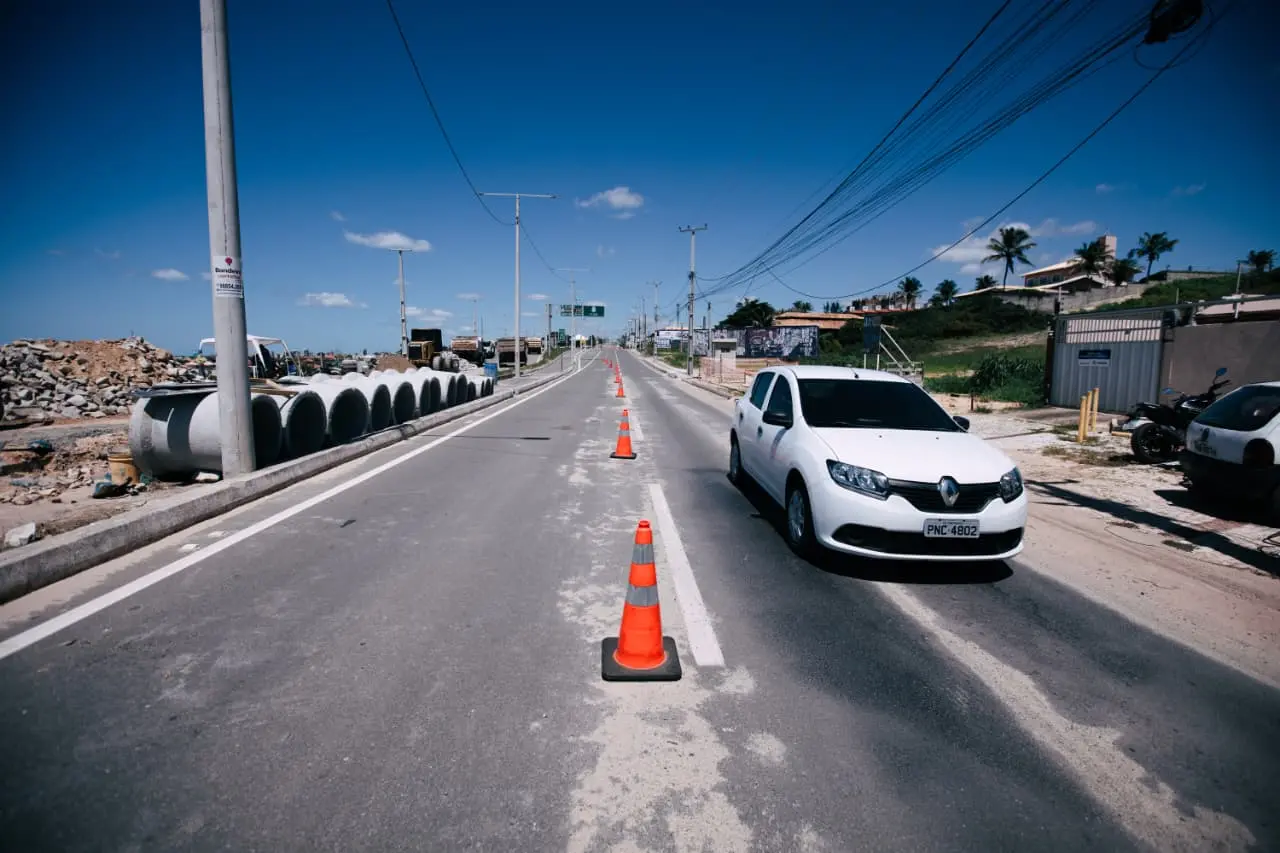 Vista de uma rodovia em obras com cones alaranjados dividindo as faixas e um carro branco circulando à direita. No canteiro esquerdo, há grandes manilhas de concreto empilhadas e postes de iluminação sob um céu azul.