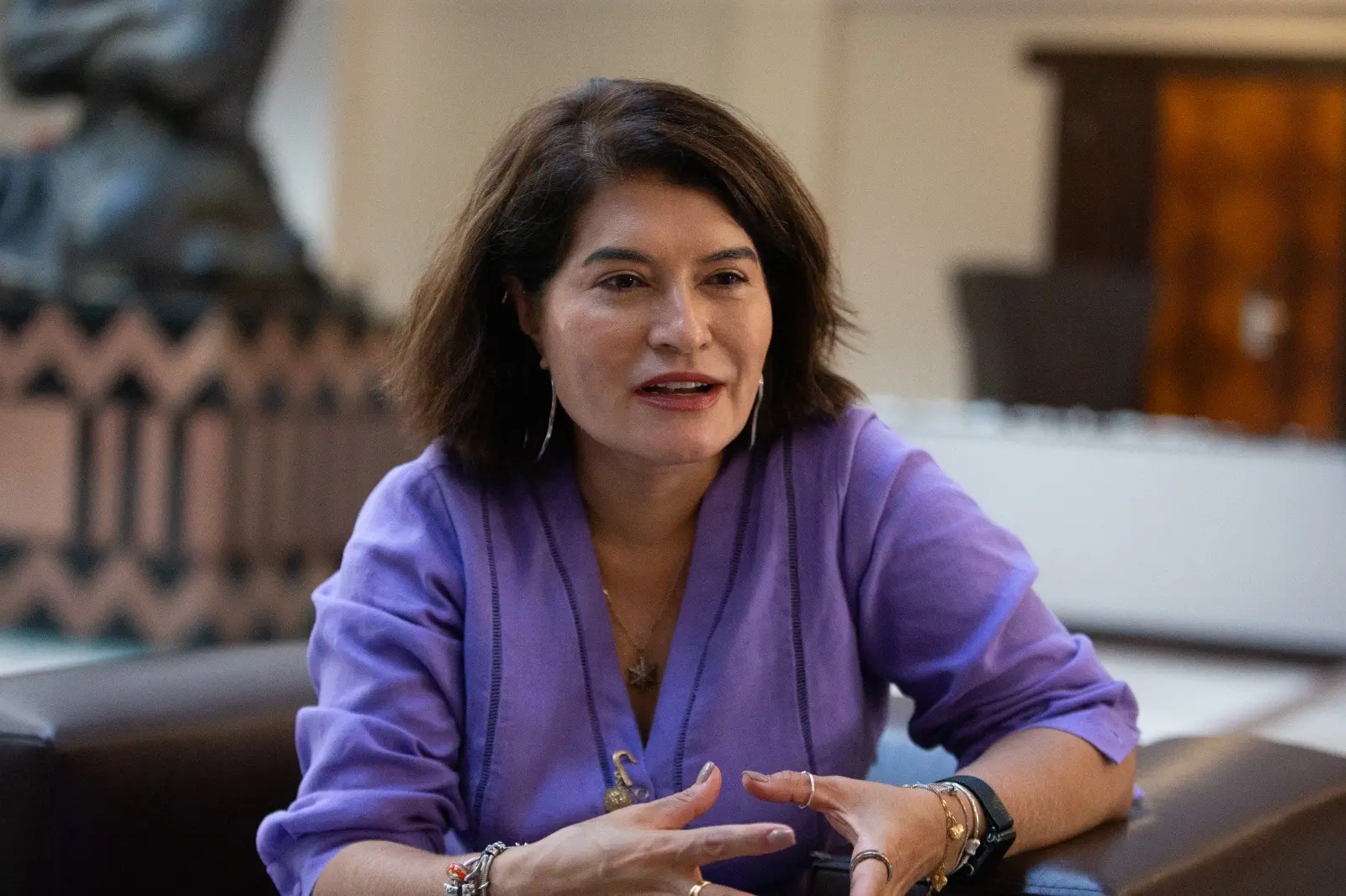 Elbia Gannoum, presidente da Abeeólica, mulher sentada em ambiente interno conversa enquanto gesticula com as mãos. Ela veste blusa roxa e acessórios, e está em um espaço com móveis e decoração ao fundo, com expressão atenta durante a fala.