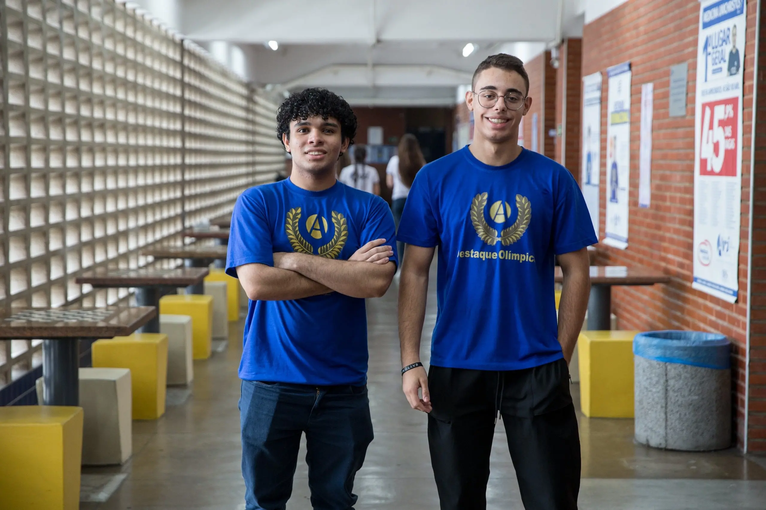 Dois jovens de camiseta azul sorriem lado a lado no corredor de uma escola. O da esquerda tem braços cruzados e o da direita usa óculos, ambos exibindo um brasão dourado no peito e a frase 