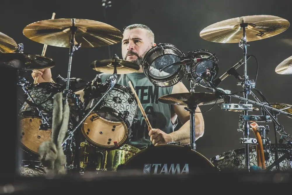 Baterista tocando bateria em um show ao vivo, com foco nas peles e tons dourados dos tambores e pratos, cenário de concertos com iluminação de palco. Perfeito para quem gosta de música ao vivo e instrumentos musicais.