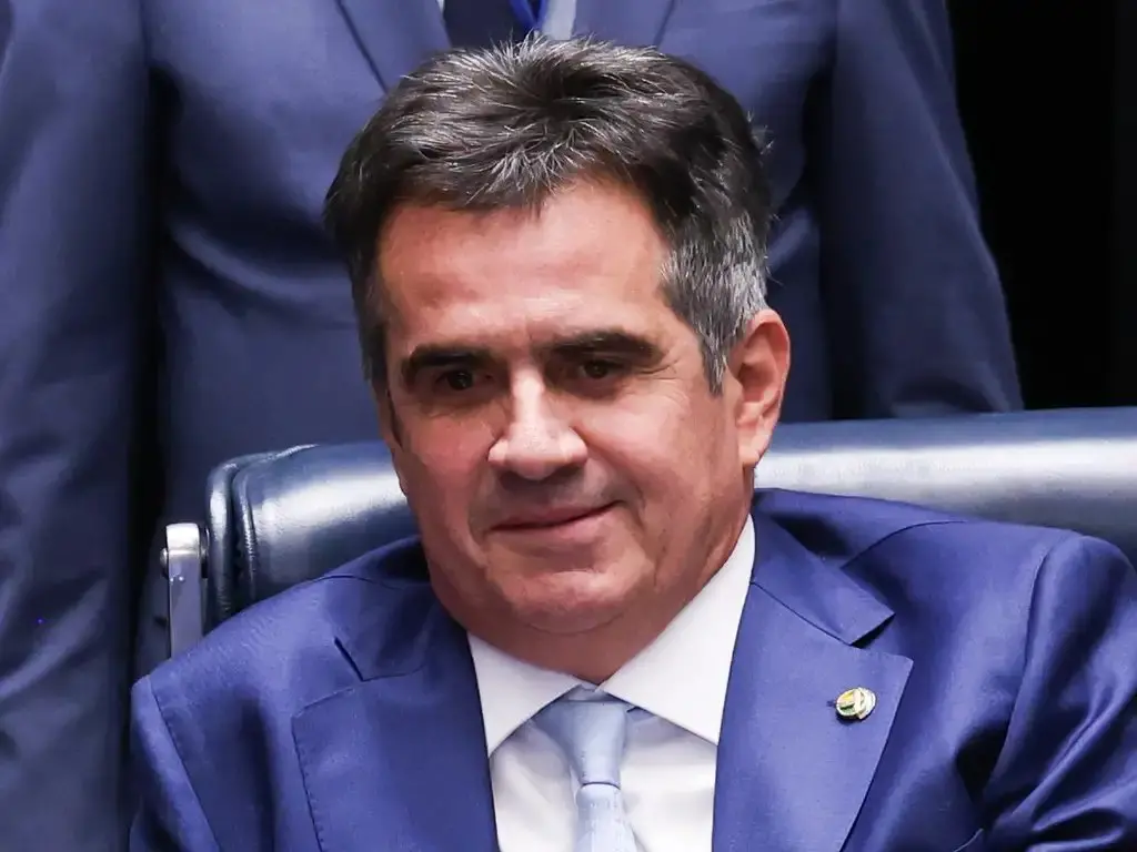 Pessoa sentada em uma cadeira, usando terno azul, camisa branca e gravata azul-clara, em ambiente interno com outras pessoas ao fundo.