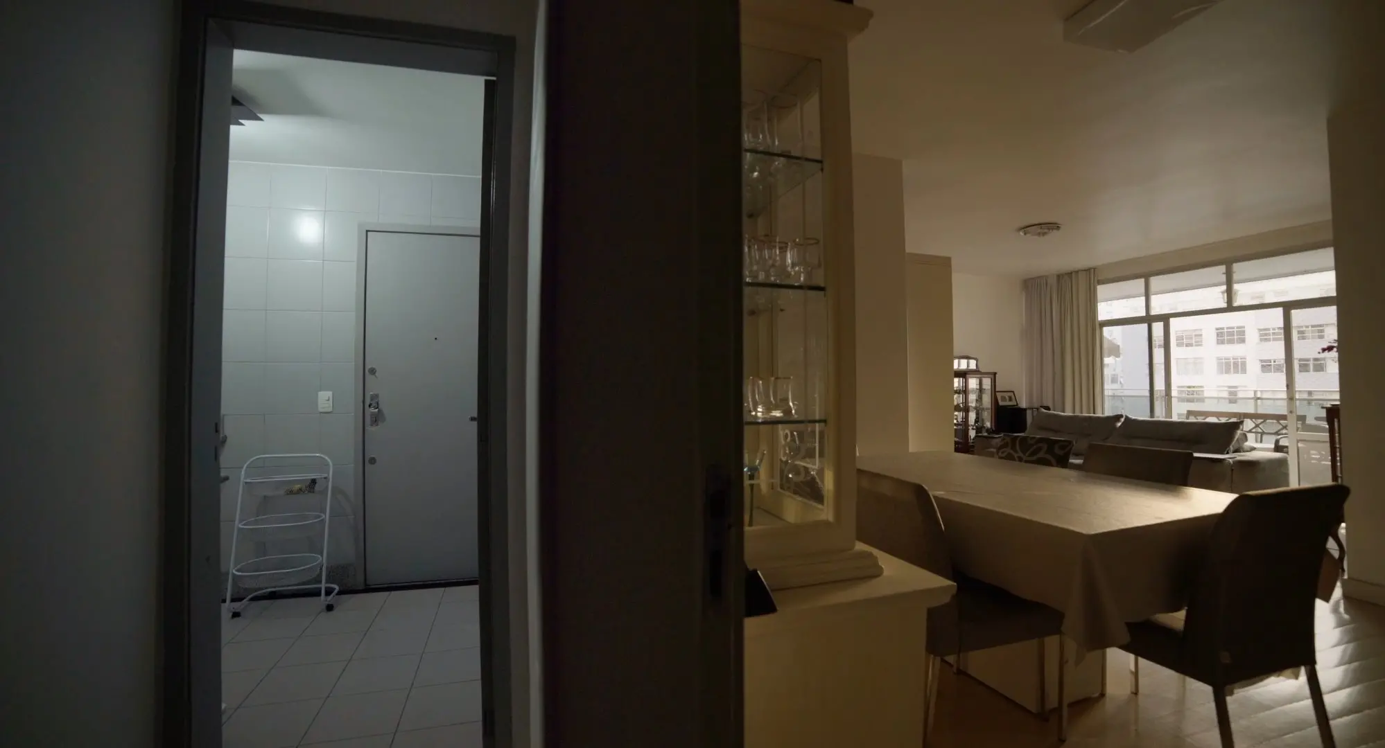 Uma vista parcial de um apartamento mostra uma área de serviço clara à esquerda e uma sala de jantar mergulhada em sombras à direita. No centro, uma divisória escura separa o ambiente iluminado por azulejos brancos do interior da sala, onde se veem uma mesa posta e uma cristaleira sob uma luz suave e filtrada.