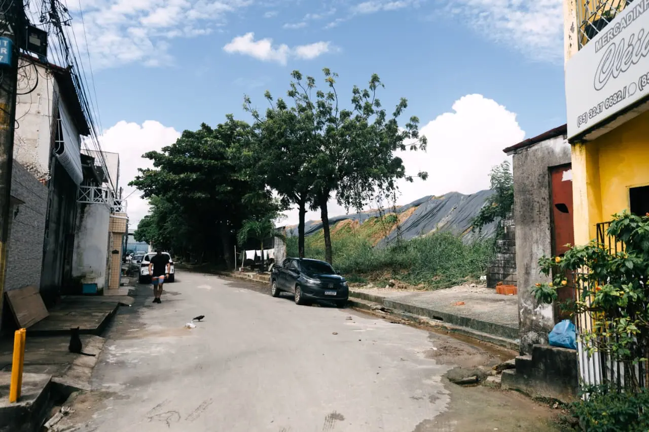 Uma rua estreita em um bairro residencial é ladeada por casas simples e árvores frondosas, enquanto um homem caminha ao fundo sob um céu azul com nuvens brancas. No lado direito, um grande aterro coberto por lonas pretas domina a paisagem, contrastando com o cotidiano urbano da vila.