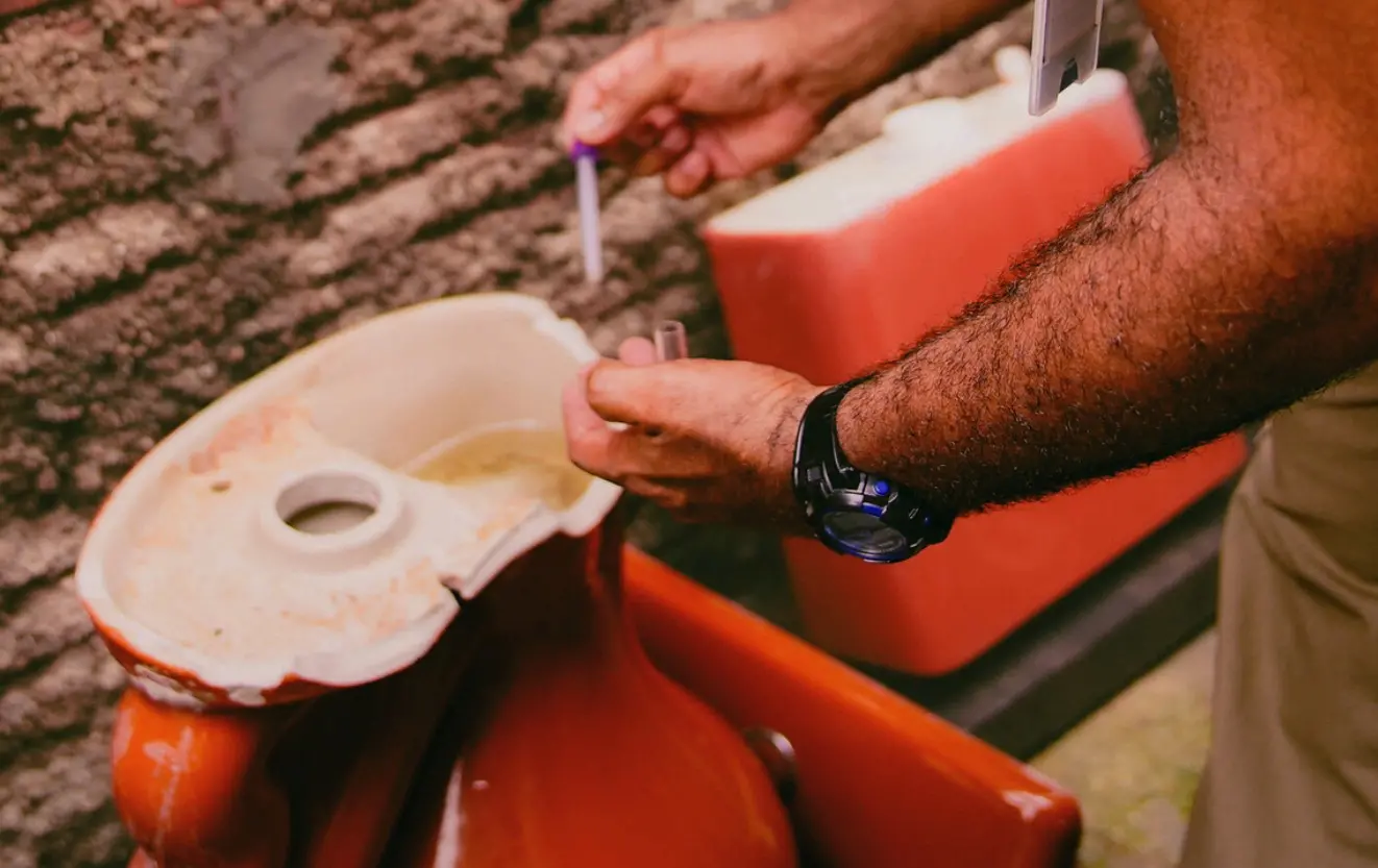 Close-up das mãos de um agente de saúde coletando amostras de água de uma caixa acoplada de descarga quebrada. Ele utiliza um conta-gotas e um tubo de ensaio para fiscalizar focos do mosquito Aedes aegypti.