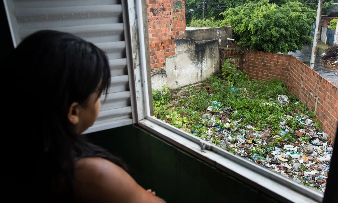 Vista de uma mulher em uma janela observando um quintal vizinho repleto de lixo, entulho e vegetação alta. O cenário ilustra um ambiente propício para o acúmulo de água e a proliferação de mosquitos transmissores de doenças.