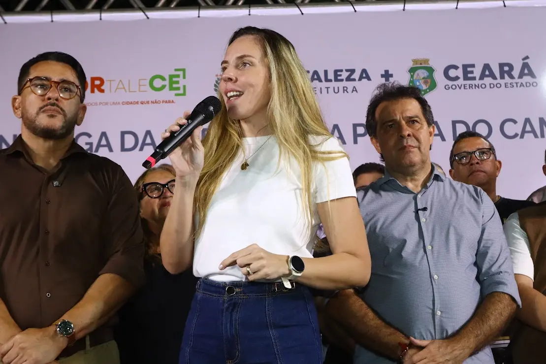 Mulher de cabelos longos e loiros fala ao microfone durante evento público em palco. Ela veste camiseta branca e calça jeans azul de cintura alta. Ao fundo, há um painel com logotipos da Prefeitura de Fortaleza e do Governo do Ceará, além de outras pessoas acompanhando o pronunciamento.