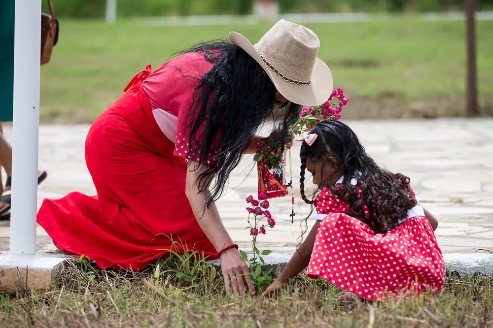 Na imagem, uma mulher e uma criança estão ajoelhadas no chão, ao lado de uma área gramada, cuidando de uma pequena muda de roseira com flores cor-de-rosa. A mulher, à esquerda, usa um chapéu de palha de abas largas, um vestido longo vermelho e segura um terço e uma pequena estampa religiosa. A criança, à direita, tem cabelos longos e cacheados com um laço rosa e veste o traje típico de devoto: um vestido vermelho com bolinhas brancas. Elas estão em um pátio de pedras claras, em um momento de aparente oração ou homenagem simbólica.