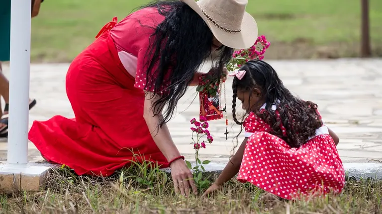 Na imagem, uma mulher e uma criança estão ajoelhadas no chão, ao lado de uma área gramada, cuidando de uma pequena muda de roseira com flores cor-de-rosa. A mulher, à esquerda, usa um chapéu de palha de abas largas, um vestido longo vermelho e segura um terço e uma pequena estampa religiosa. A criança, à direita, tem cabelos longos e cacheados com um laço rosa e veste o traje típico de devoto: um vestido vermelho com bolinhas brancas. Elas estão em um pátio de pedras claras, em um momento de aparente oração ou homenagem simbólica.