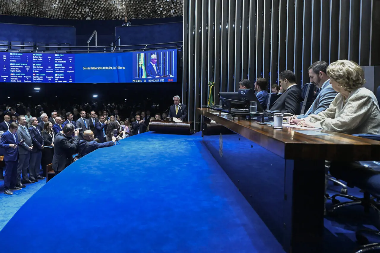 Interior de um plenário, com chão azul, políticos ao fundo e mesa principal preenchida.