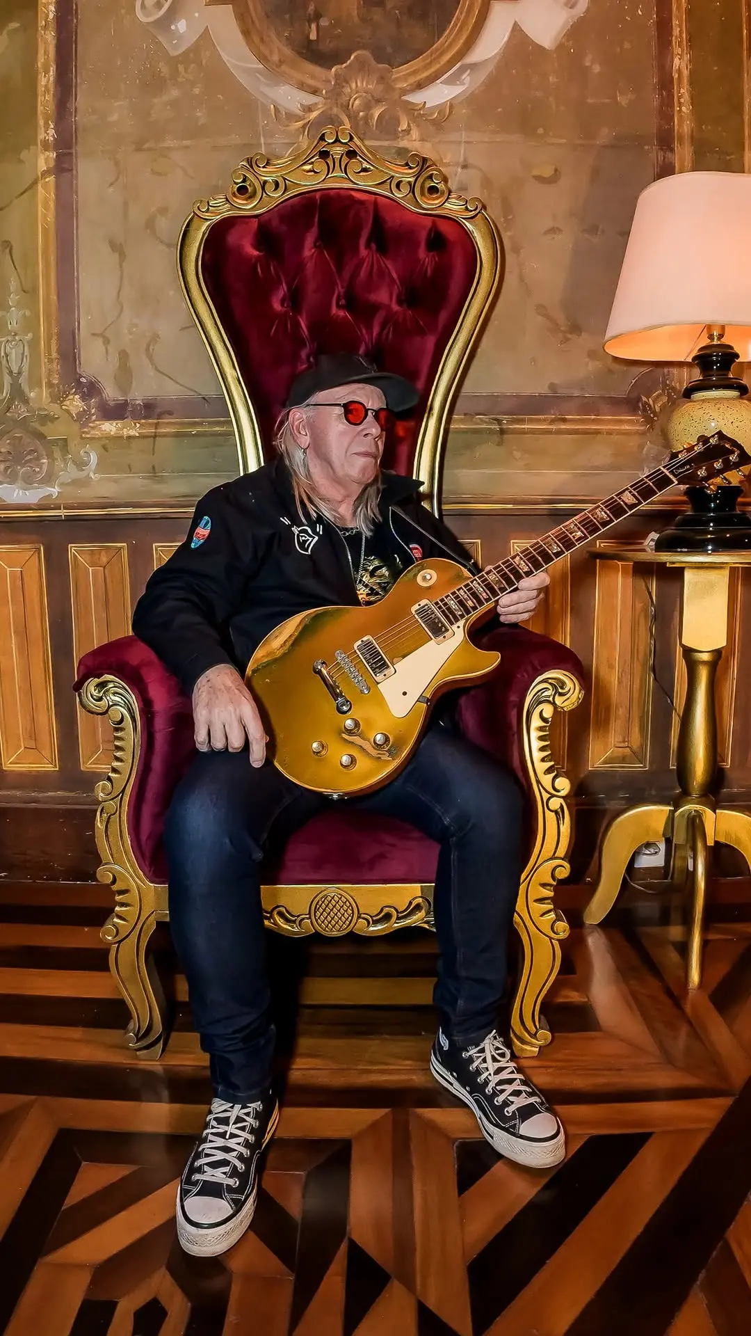 Homem jovem com óculos vermelhos, cabelo grisalho, usando boné preto, vestido casual e tênis, tocando guitarra elétrica dourada em um ambiente luxuoso com cadeira adornada e iluminação quente.
