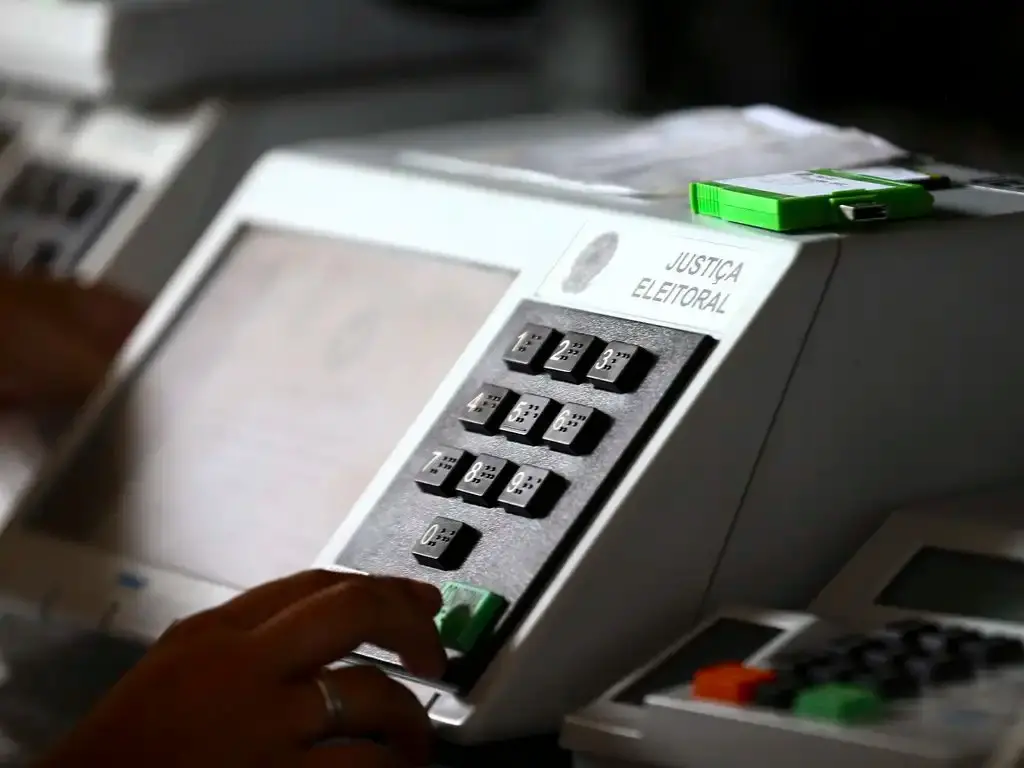 Urna eletrônica com dígitos de 0 a 9 e botão verde, com dedo em cima.