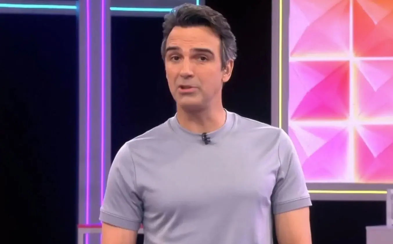 Pessoa com camiseta cinza fala em estúdio de televisão, com fundo iluminado por luzes coloridas e painéis geométricos.