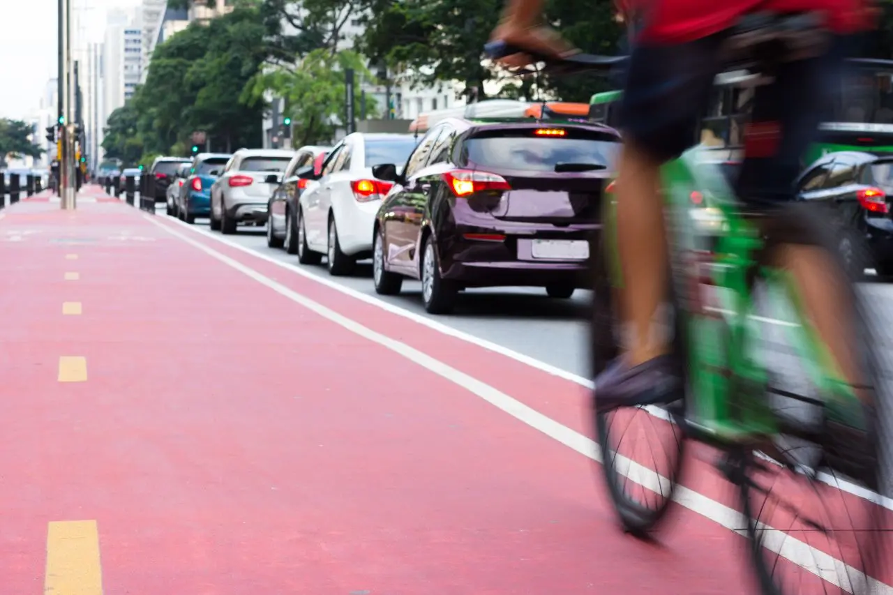 Ciclovia com bicicleta verde borrada passando, com fila de carros ao fundo em uma via urbana.