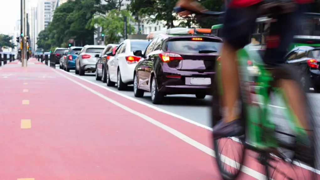 Ciclovia com bicicleta verde borrada passando, com fila de carros ao fundo em uma via urbana.