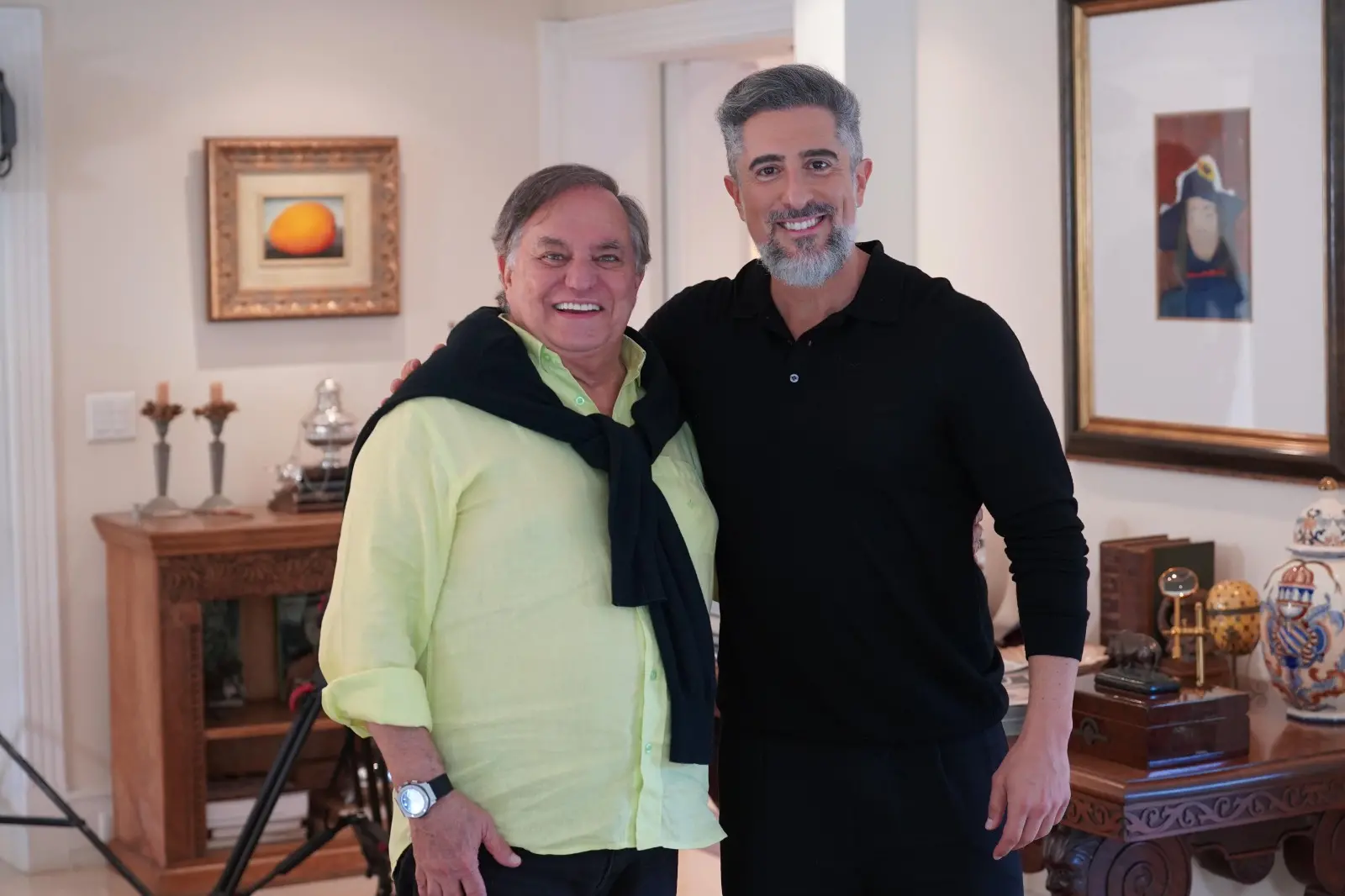 Dois homens sorridentes em uma casa elegante, um com blazer amarelo e o outro com blusa preta, comemorando um momento de felicidade juntos.