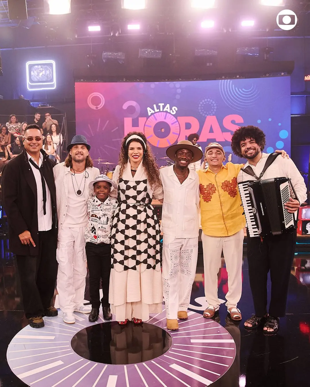 Grupo de músicos e performers em palco com iluminação artística e tela de fundo exibindo 'Altas Horas' em destaque, todos sorrindo para a câmera.