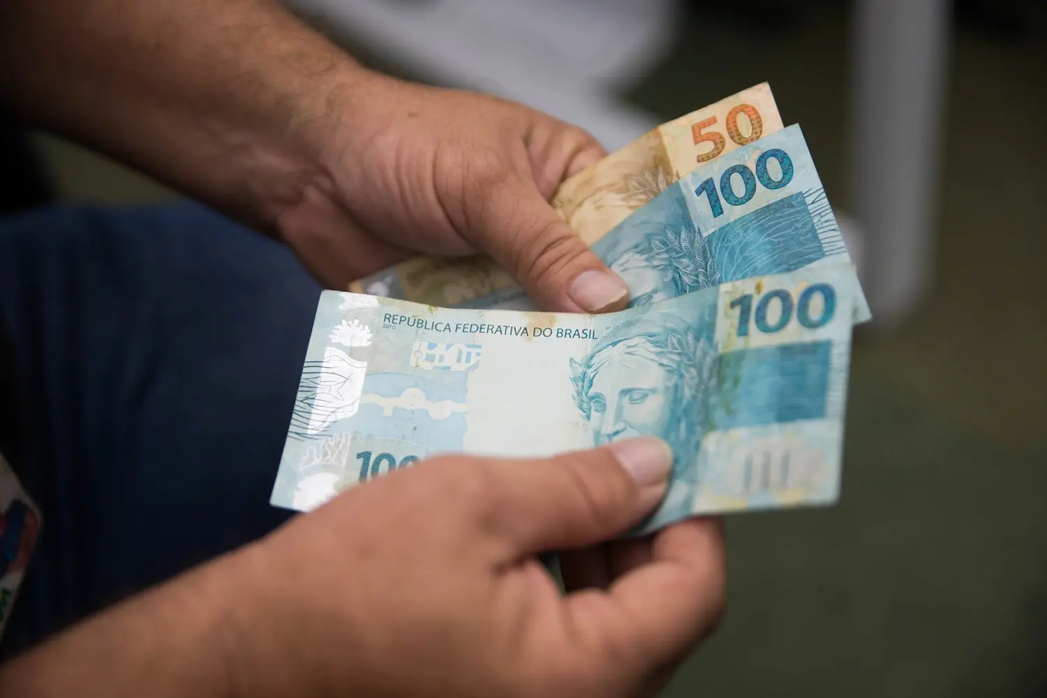 Mãos seguram notas de Real brasileiro em primeiro plano, destacando cédulas de cem e de cinquenta reais sobre uma superfície clara. A imagem foca no manuseio do dinheiro, apresentando os detalhes das cores azul e laranja das notas em uma composição nítida e direta.