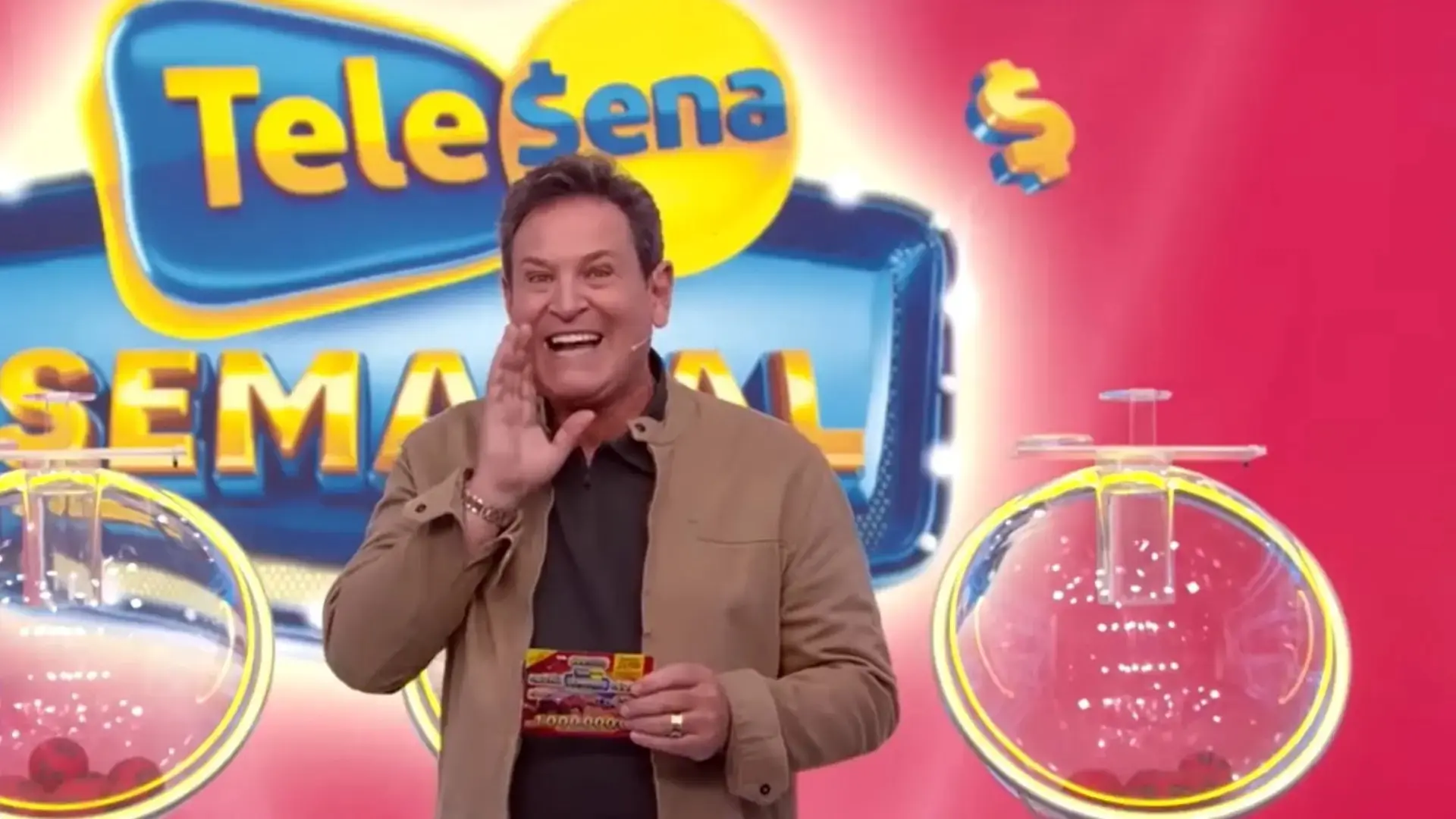 O apresentador sorri e faz um gesto de anúncio no palco da 'Tele Sena Semanal', destacando o título de capitalização em suas mãos enquanto globos de sorteio ladeiam a cena. O cenário vibrante em tons de rosa e azul compõe a atmosfera festiva da premiação em uma composição dinâmica e colorida.