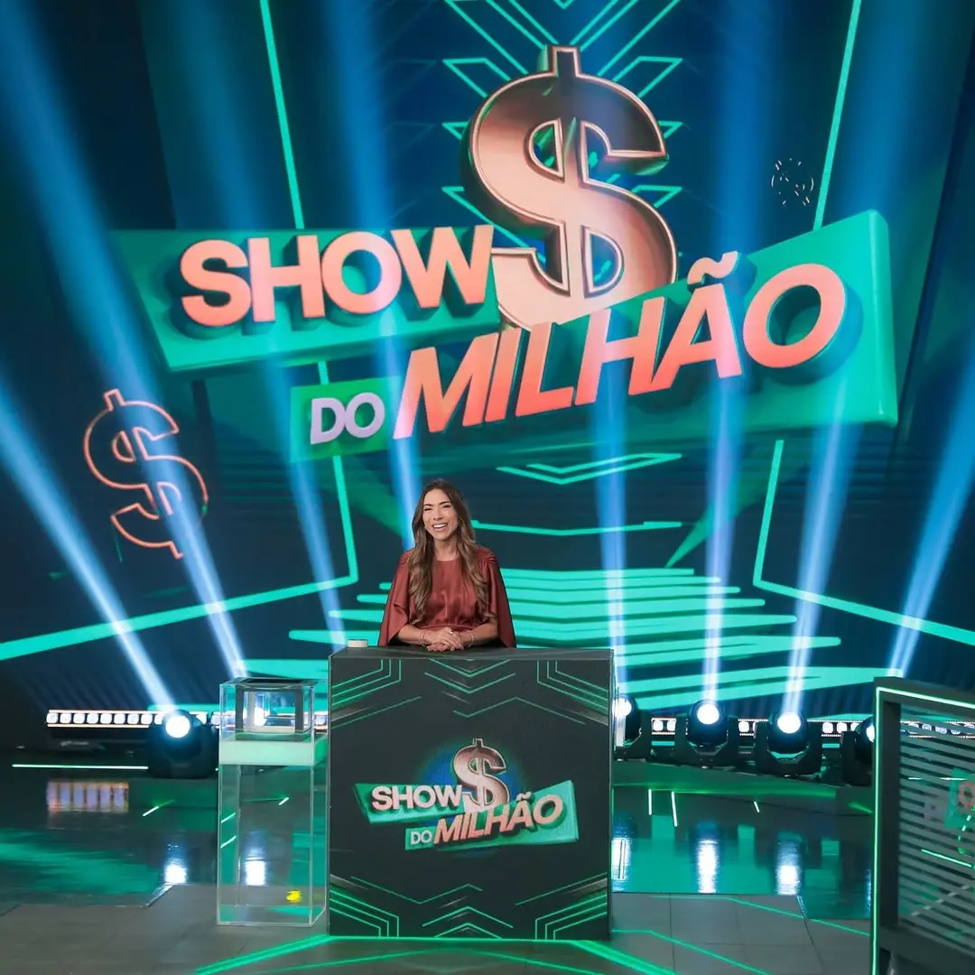 A apresentadora Patricia Abravanel sorri atrás de um púlpito no centro do palco do 'Show do Milhão', destacando o logotipo da atração sob um grande cifrão dourado ao fundo. O cenário é composto por feixes de luz neon verdes e azuis que criam uma atmosfera tecnológica e vibrante em uma composição de cores intensas.