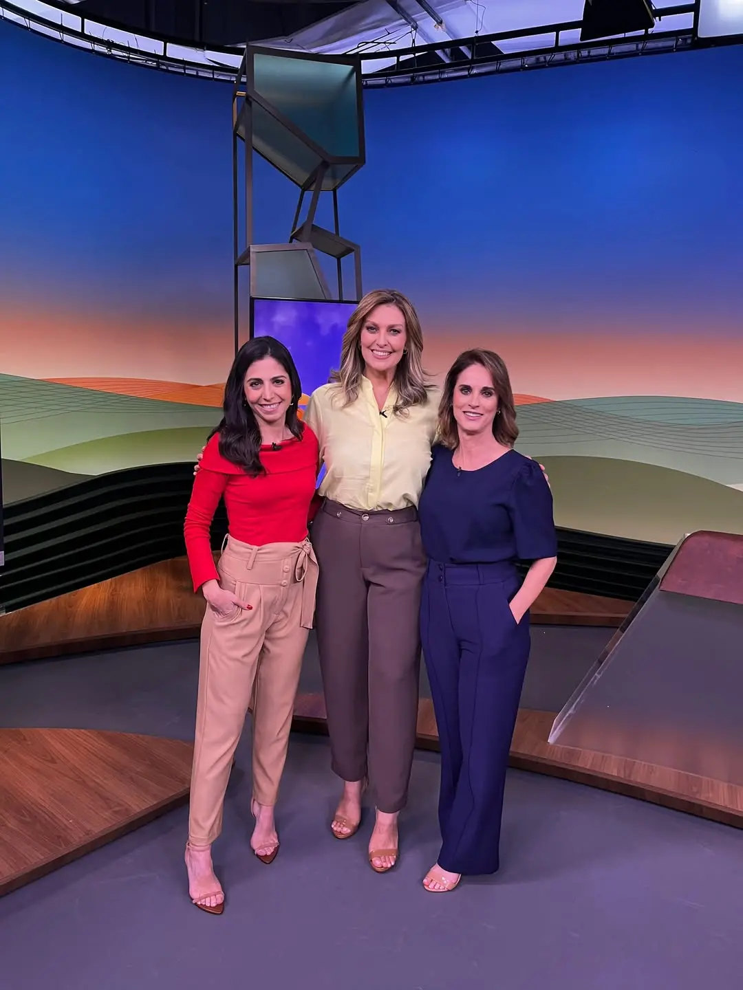 Três apresentadoras em pé, lado a lado, em um estúdio de televisão, com cenário colorido e bancada ao fundo.