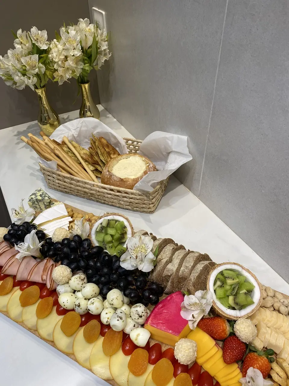 Mesa com tábua de frios e frutas organizada com uvas, morangos, kiwi, maçã, damasco, queijos, pães e embutidos. Ao fundo, cesta com torradas e creme servido no pão, além de vasos dourados com flores brancas decorando o ambiente.