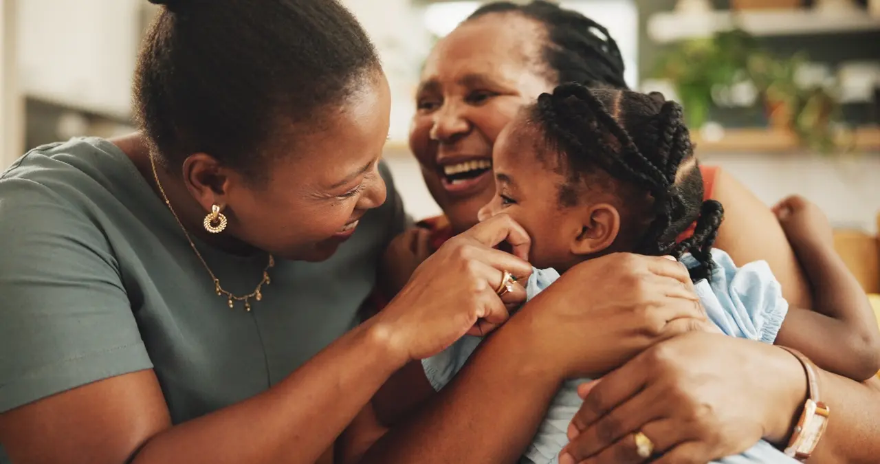 Três gerações de uma família negra compartilham um momento de afeto, com duas mulheres sorrindo enquanto abraçam e brincam com uma criança de tranças. A cena transmite calor e união por meio de expressões alegres e luz suave em um ambiente doméstico acolhedor.