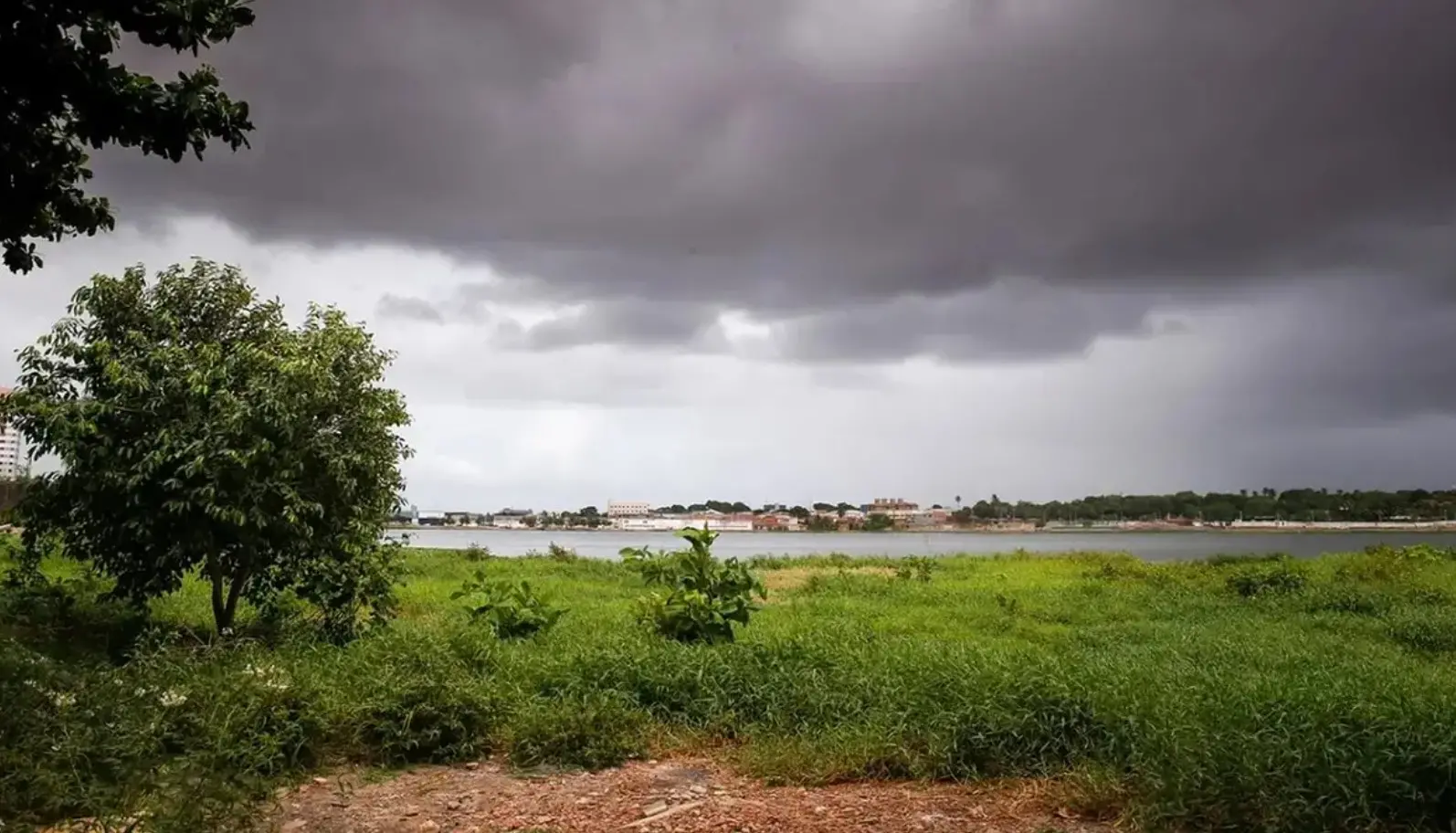 Céu encoberto por nuvens escuras sobre área verde às margens de um lago, com vegetação no primeiro plano e construções ao fundo, indicando tempo chuvoso no Ceará.
