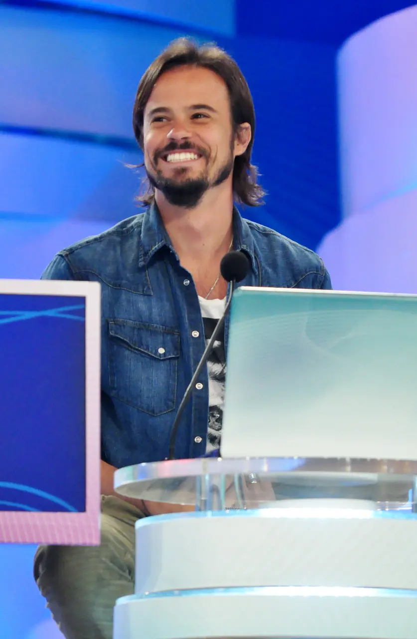 Homem sorrindo em estúdio de televisão, usando camisa jeans azul sobre camiseta branca, sentado atrás de uma bancada com microfone e monitores, com iluminação em tons de azul ao fundo.
