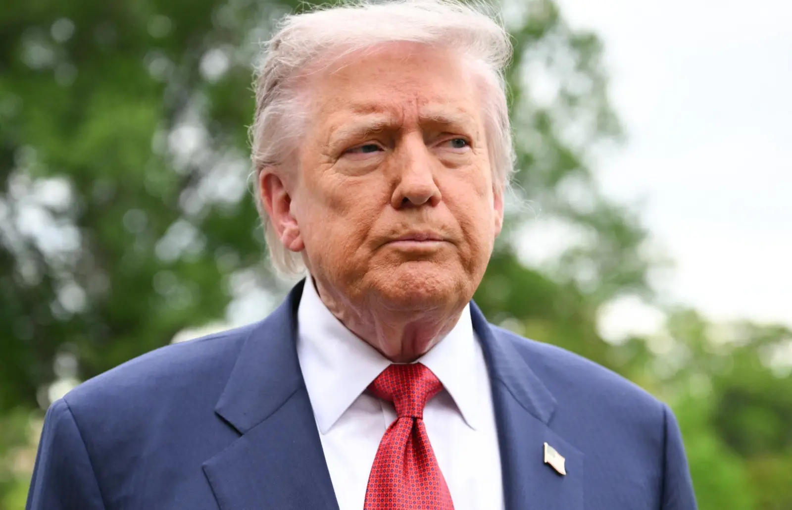Donald Trump é um homem idoso, branco, de cabelo liso branco. Na foto, ele usa terno azul, camisa social branca, gravata vermelha e está com uma expressão séria.
