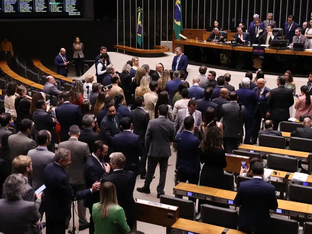Imagem aérea interna de um plenário, com mesas de madeira e diversos parlamentares em pé, trajando roupas formais predominantemente azul marinho.