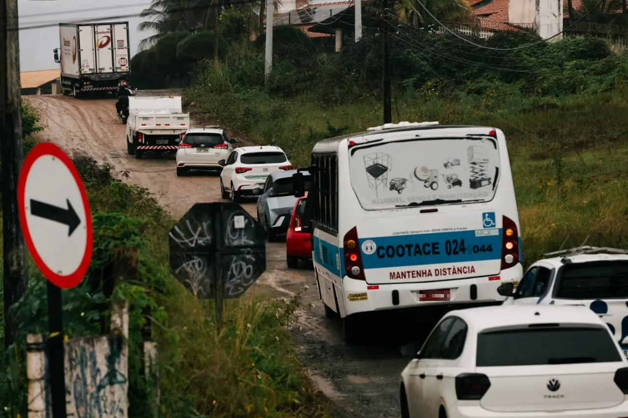 Fila de veículos, incluindo carros de passeio, um ônibus da COOTACE e um caminhão, trafegando por um desvio de terra estreito e irregular em um dia nublado. À esquerda, uma placa circular branca com borda vermelha e seta preta indica a direção do fluxo, cercada por vegetação alta e postes de luz.