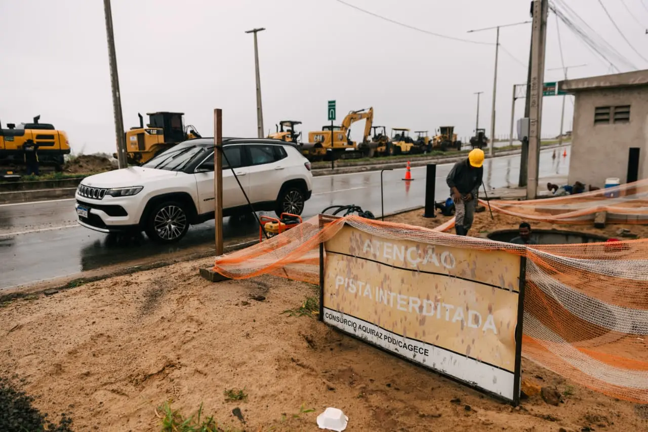 Placa amarela desgastada com o aviso ATENÇÃO PISTA INTERDITADA em destaque, posicionada em um canteiro de obras de terra sob chuva fina. Ao fundo, máquinas pesadas amarelas estão estacionadas ao longo de uma rodovia asfaltada onde um SUV branco transita, enquanto operários trabalham em uma abertura no solo.