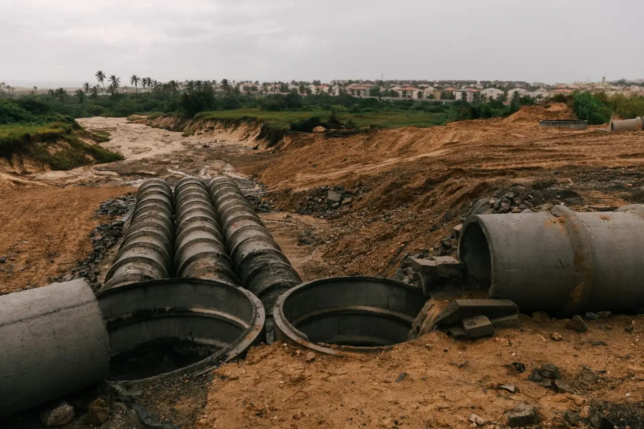 Canteiro de obras de drenagem apresentando grandes manilhas de concreto alinhadas e outras parcialmente soterradas em um terreno de terra batida e lama. A paisagem ao fundo revela uma área de vale com erosão, vegetação rasteira e um conjunto de casas brancas sob um céu cinzento e carregado.