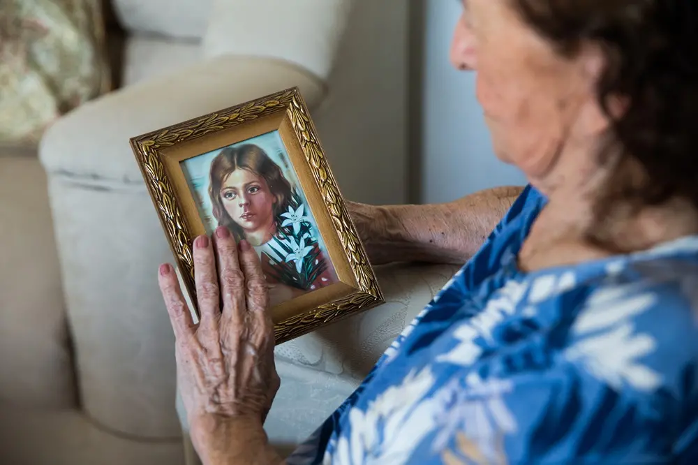 Na imagem, uma mulher idosa, vista de perfil, segura com as duas mãos um pequeno porta-retrato dourado e ornamentado. Dentro da moldura, há uma ilustração de uma jovem de cabelos castanhos (Beata Benigna), que segura flores brancas (lírios) contra o peito. A mão esquerda da idosa repousa delicadamente sobre o vidro da imagem. O fundo está desfocado, mostrando parte de um sofá claro e a roupa azul estampada da mulher.