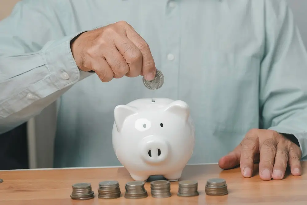 Pessoa colocando uma moeda em um cofrinho de porquinho branco, simbolizando economia, finanças pessoais ou investimento inteligente.