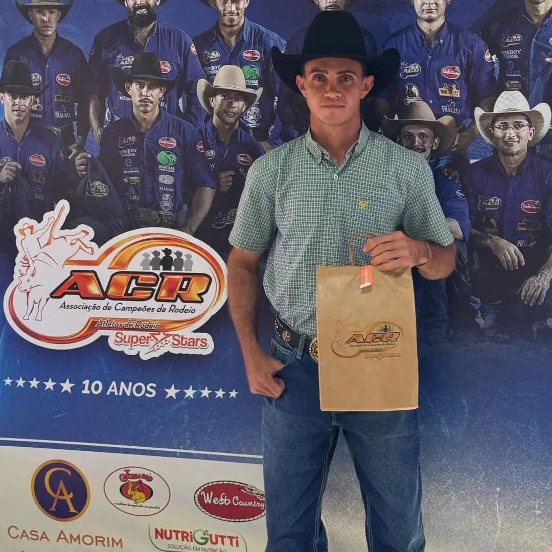 Um jovem peão de rodeio posa em pé vestindo chapéu preto, camisa xadrez verde e calça jeans enquanto segura uma sacola de papel pardo. Ao fundo, um painel azul exibe a marca da Associação de Campeões de Rodeio (ACR) e fotografias de outros competidores em destaque.