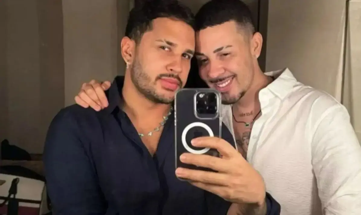 Carlinhos Maia e seu ex-esposa posam juntas para uma selfie em frente ao espelho, usando roupas de cores escura e clara; uma delas segura um celular, e o ambiente aparenta ser um espaço interno com iluminação artificial. Imagem usada em matéria sobre liminar pedido pelo motorista contra o influenciador.