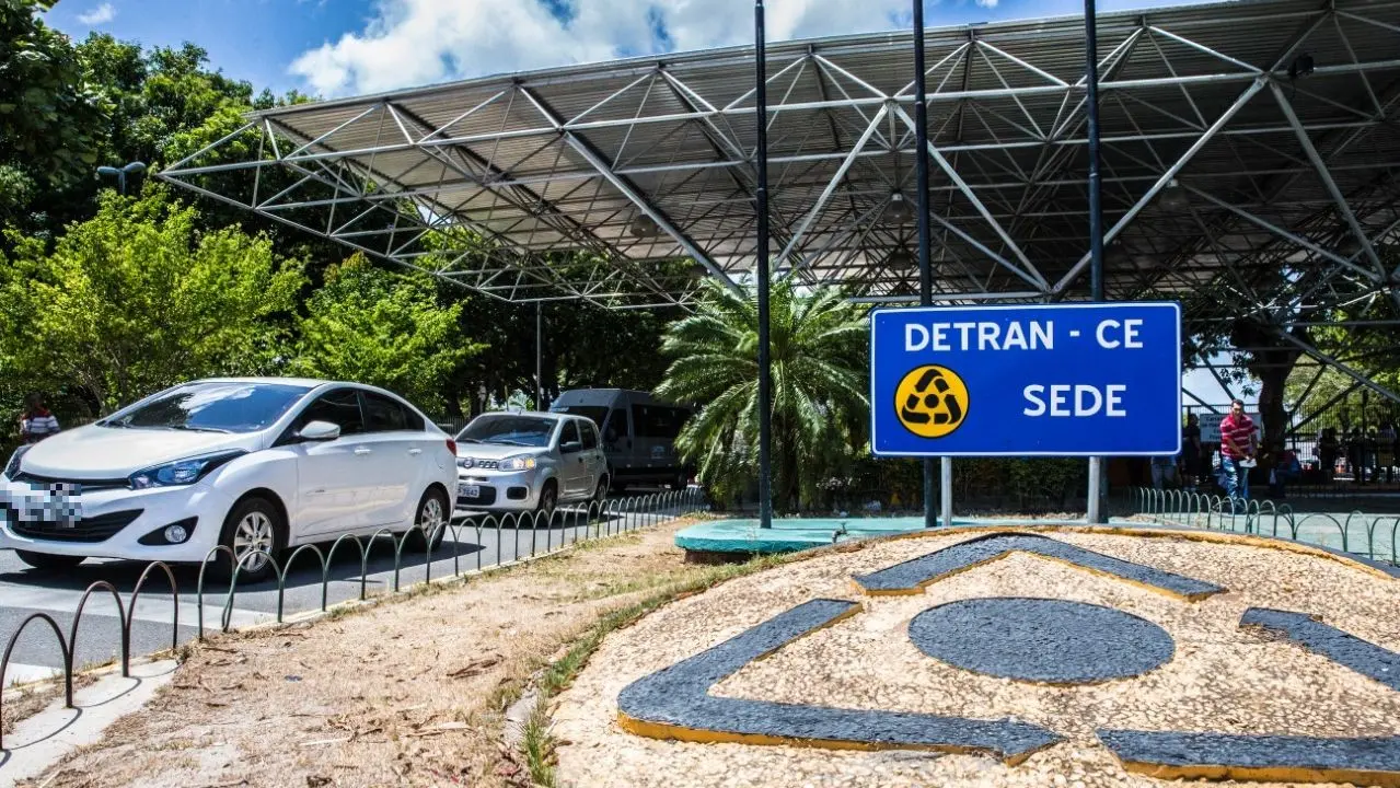 Fachada da sede do Detran Ceará, com placa azul “DETRAN‑CE Sede” em primeiro plano, estrutura coberta ao fundo e carros passando pela via ao lado, em área arborizada.
